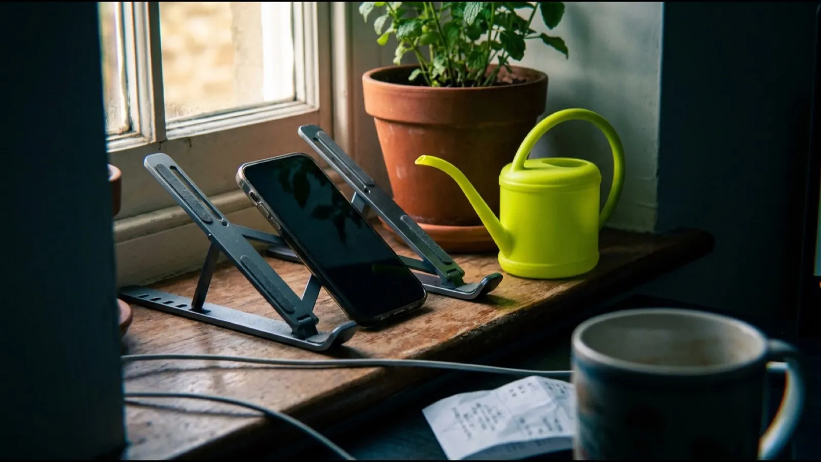 A phone propped against a laptop stand on a windowsill beside a plant
