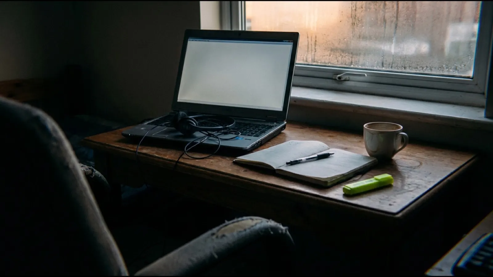 A laptop open on a desk at dusk with a headset resting on the keyboard